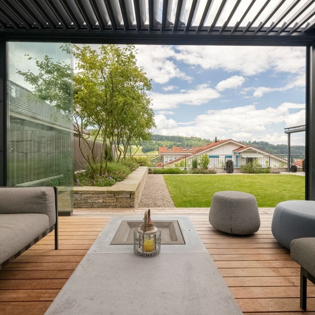 Blick aus einem SKL 100 Lamellendach mit Glaswänden in Malters, Luzern – moderner Outdoor-Bereich mit Lounge-Möbeln, Holzdeck und Blick ins Grüne.