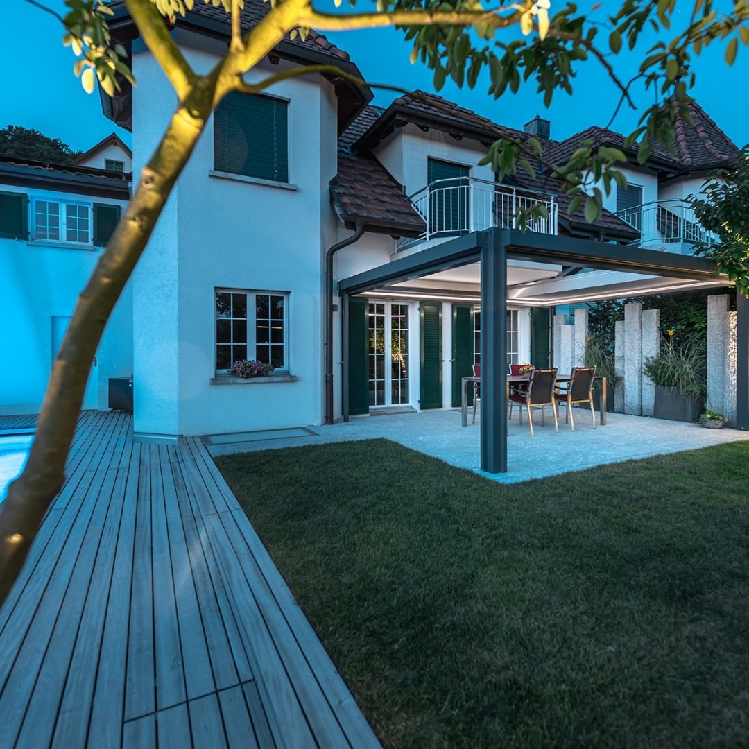 SKL 150 Panorama Plus Lamellendach bei Abendstimmung mit LED-Beleuchtung, vollständig zurückgefahrenen Lamellen und gedecktem Essbereich im Garten.