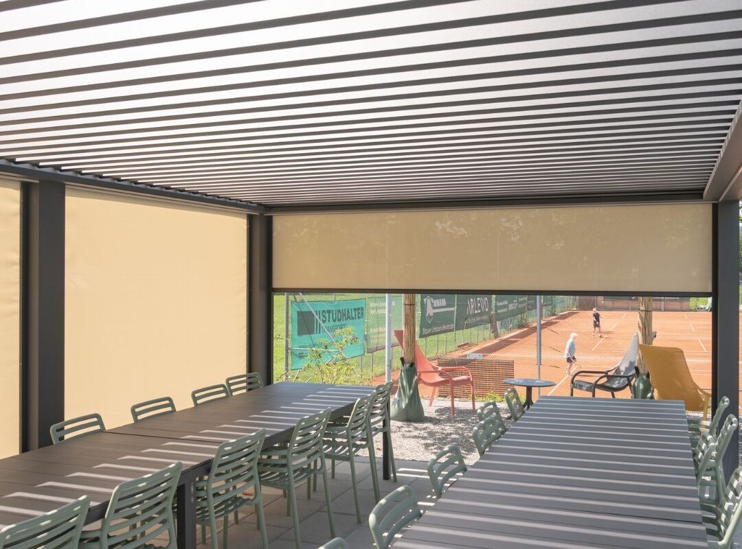 Innenansicht des Lamellendachs beim Tennisclub Horw in Luzern mit geschlossenen Seitenmarkisen, langen Tischen und Blick auf die Tennisplätze.