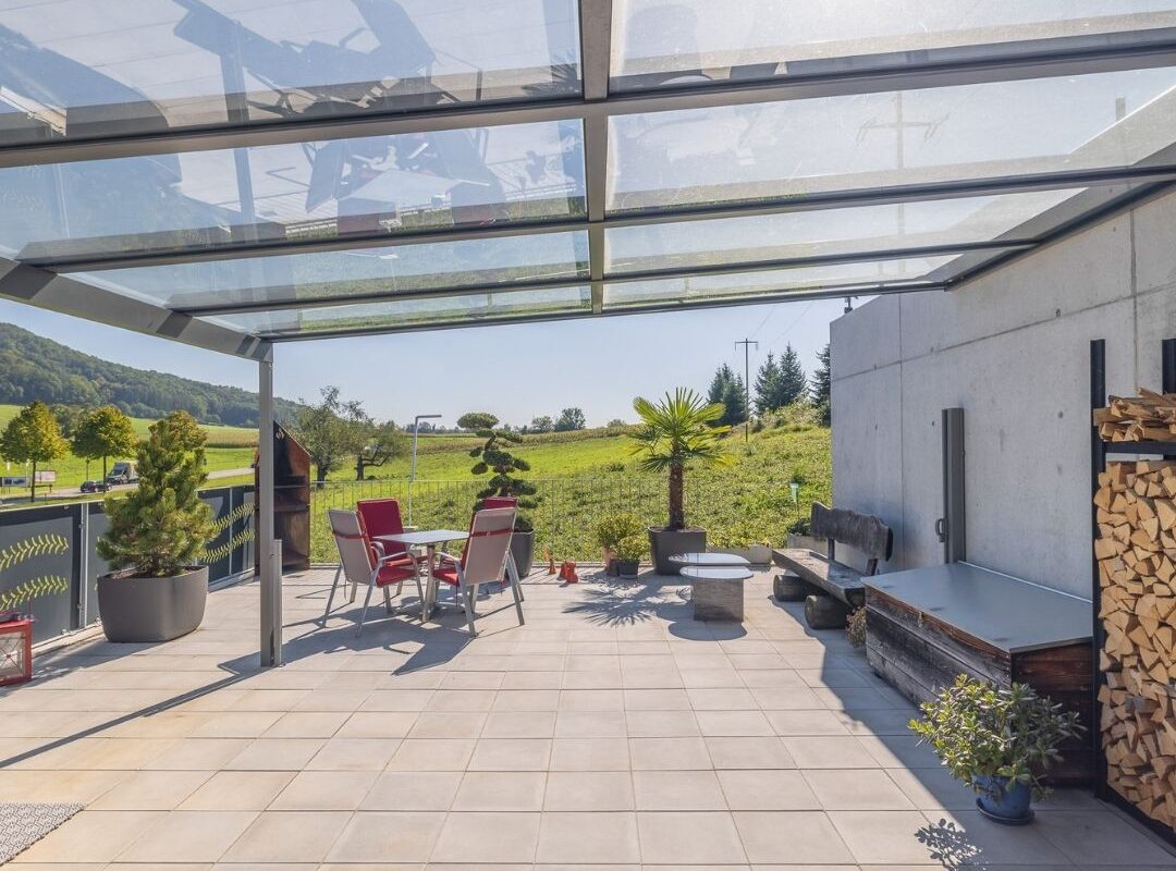 Grosszügige Terrasse mit Glasdach, modernen Gartenmöbeln und Blick ins Grüne – lichtdurchfluteter Outdoor-Bereich mit natürlichem Ambiente.