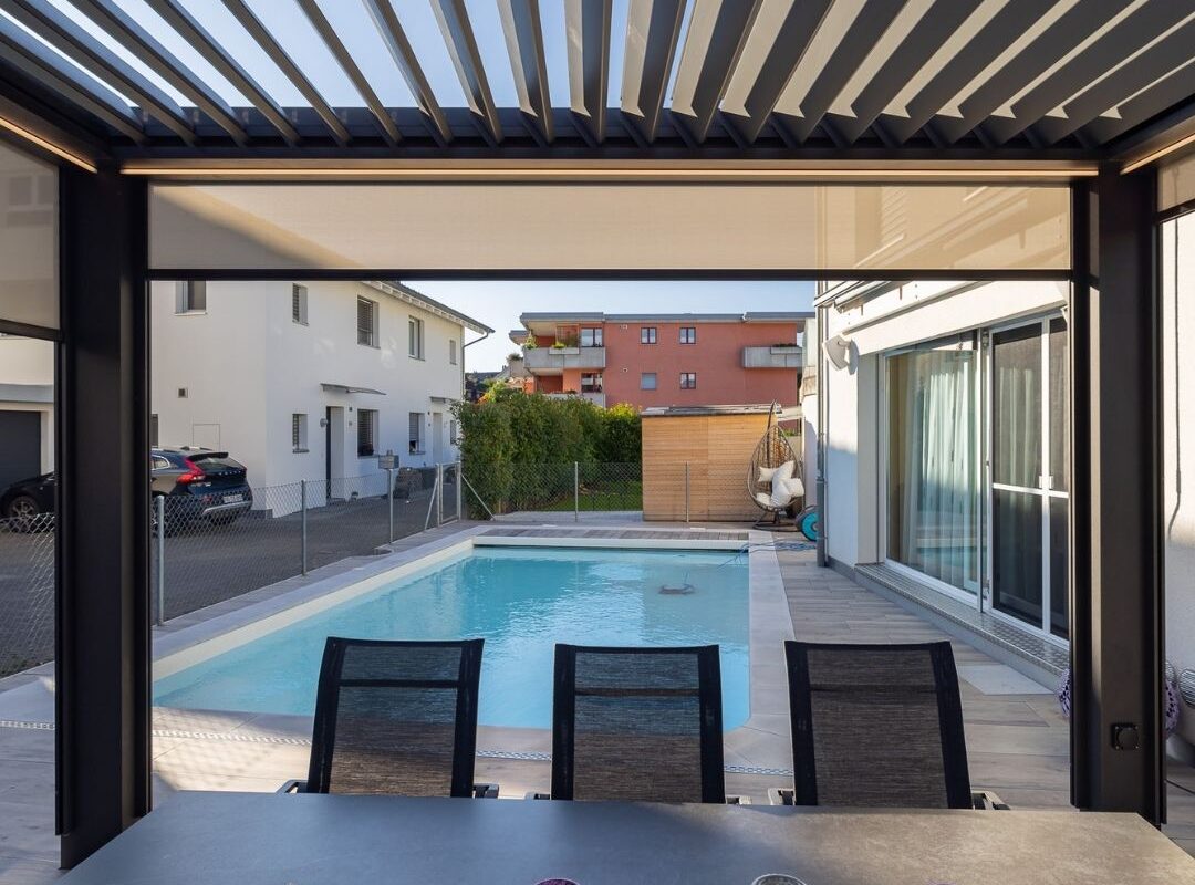 Blick vom gedeckten Essbereich unter einem Lamellendach auf den Pool im Garten.