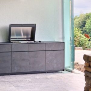Avers Outdoor-Küche mit geöffnetem Grillmodul und eleganten dunklen Fronten in Steinoptik, platziert in einem modernen Outdoorbereich mit Glaswänden.