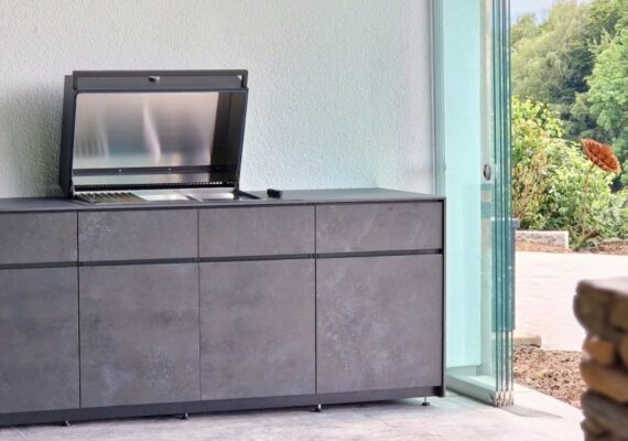 Avers Outdoor-Küche mit geöffnetem Grillmodul und eleganten dunklen Fronten in Steinoptik, platziert in einem modernen Outdoorbereich mit Glaswänden.