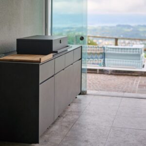 Moderne Avers Outdoor-Küche in dunkler Steinoptik mit Grillmodul und Holzarbeitsfläche, platziert in einem überdachten Outdoorbereich mit Blick auf die Landschaft.