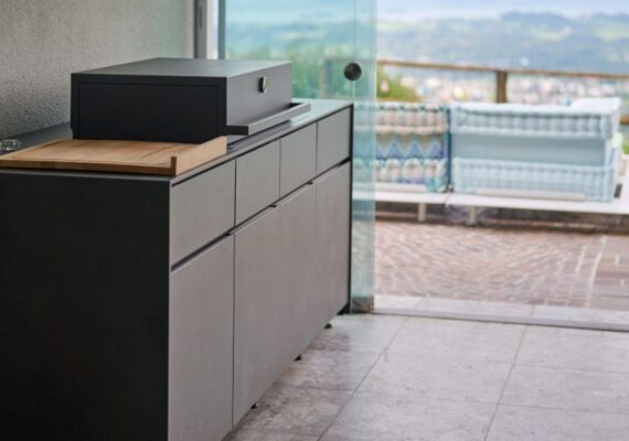 Moderne Avers Outdoor-Küche in dunkler Steinoptik mit Grillmodul und Holzarbeitsfläche, platziert in einem überdachten Outdoorbereich mit Blick auf die Landschaft.