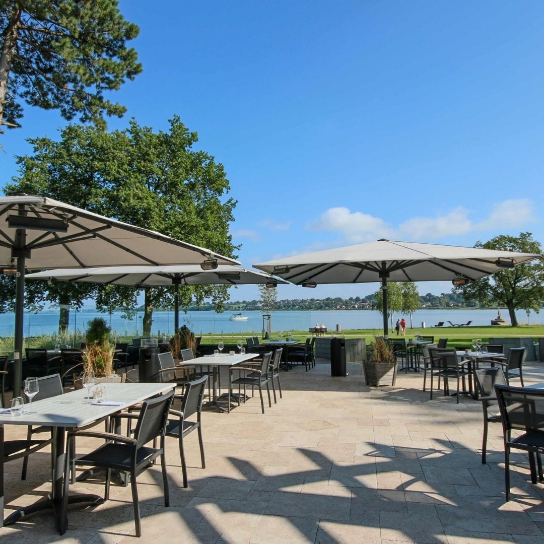 Elegante ALBATROS Grossschirme in Grau überdachen die Terrasse eines Restaurants direkt am See und bieten Gästen grosszügigen Schatten bei traumhafter Aussicht.