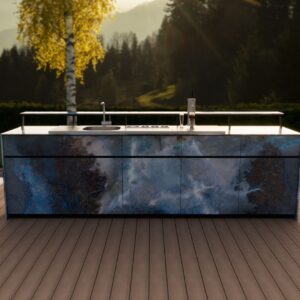 Theiss Outdoor-Bar mit blauen Steinfronten, Spülbecken und Zapfanlage auf Terrasse im Abendlicht mit Wald- und Bergkulisse.