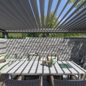 Detailaufnahme einer Pergola mit Lamellendach, die durch ihr Schattenspiel auf Tisch und Steinmauer eine architektonische Tiefe erzeugt.