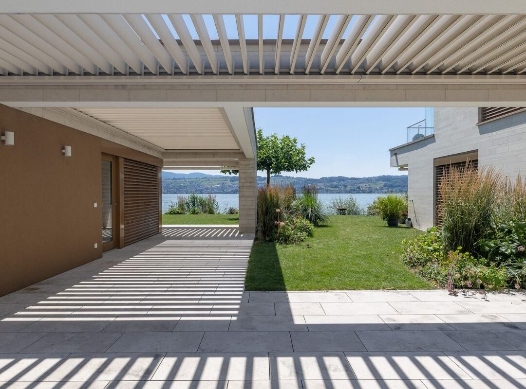 Moderner Neubau mit integriertem Lamellendach, das den Übergang von Wohnraum und Garten elegant überdacht und zugleich einen Blick auf den See freigibt.