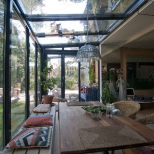 Wintergarten mit Glasdach und Glaswänden, eingerichtet mit Holztisch, Pflanzen und gemütlicher Dekoration.