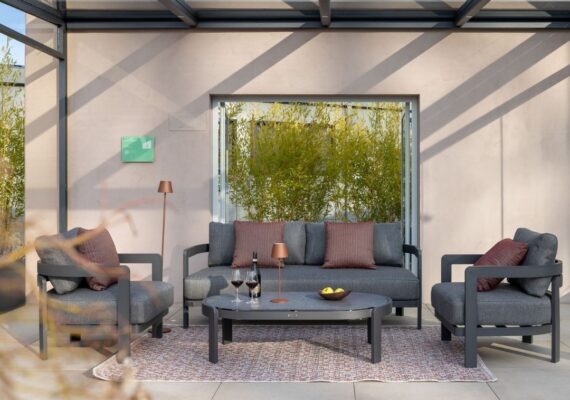 Elegante Nook Outdoor-Lounge in Grau mit passenden Sesseln, Couchtisch und Accessoires unter einem Glasdach.