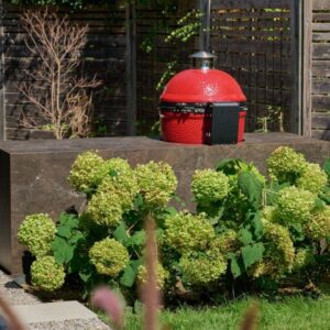 Roter Keramikgrill auf einer Outdoor Küche aus Naturstein mit blühenden Hortensien im Vordergrund.