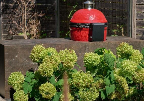 Roter Keramikgrill auf einer Outdoor Küche aus Naturstein mit blühenden Hortensien im Vordergrund.