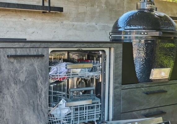Outdoor Küche im dunklen Stein-Look mit integriertem Keramikgrill und geöffneter Geschirrspülmaschine.