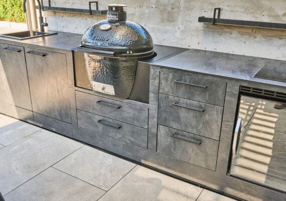 Outdoor Küche im dunklen Stein-Look mit integriertem Keramikgrill, Spülbecken und Kühlschrank.