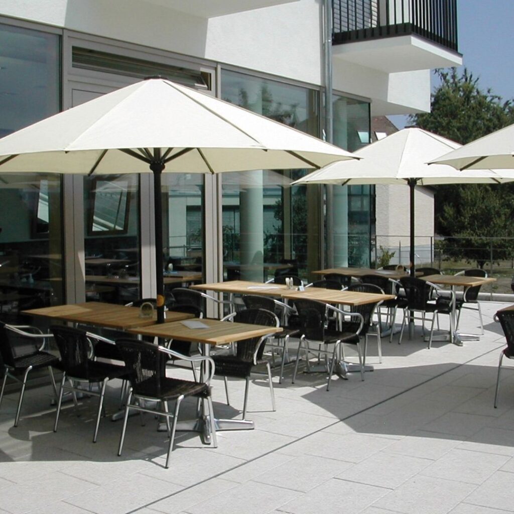 Grosse weisse Sonnenschirme auf einer modernen Gastronomie-Terrasse mit mehreren Sitzgruppen.