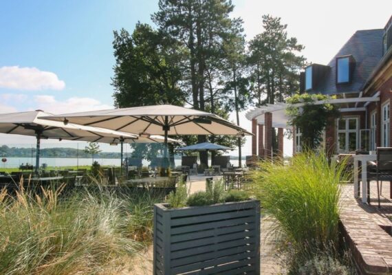 Mehrere grossflächige Sonnenschirme spenden Schatten auf einer Restaurantterrasse mit Blick auf den See.