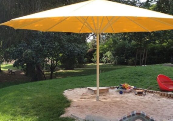 Gelber Sonnenschirm spendet Schatten über einem Sandkasten auf einer Grünfläche.