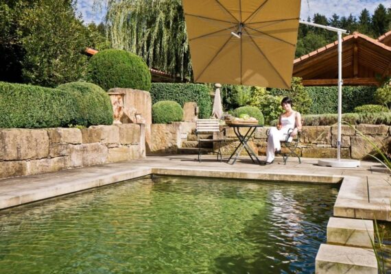 Moderner Sonnenschirm in Gelb spendet Schatten über einem Naturpool mit Sitzgelegenheit und stilvoller Gartengestaltung.
