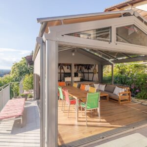 Moderner Wintergarten mit Glasdach und Beschattung, ausgestattet mit bunten Stühlen und einem grossen Holztisch, offene Bauweise mit Blick ins Grüne.