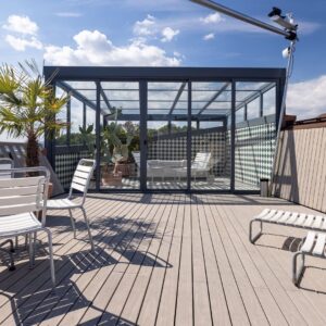 Moderner Wintergarten mit Glaswänden und Glasdach auf einer Dachterrasse mit Holzdeck und Palmen.