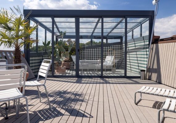 Moderner Wintergarten mit Glaswänden und Glasdach auf einer Dachterrasse mit Holzdeck und Palmen.