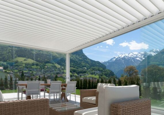 Weisses Lamellendach mit Glaswänden und moderner Garteneinrichtung, Blick auf eine Berglandschaft mit schneebedeckten Gipfeln.