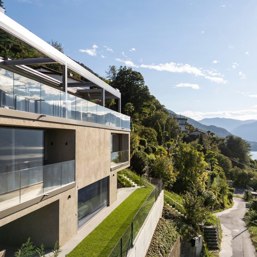 Seitliche Ansicht eines privaten Wohnhauses in Gerra im Tessin mit Lamellendach auf der Terrasse, Glasbrüstung und freiem Blick in die Hügellandschaft und Richtung See.