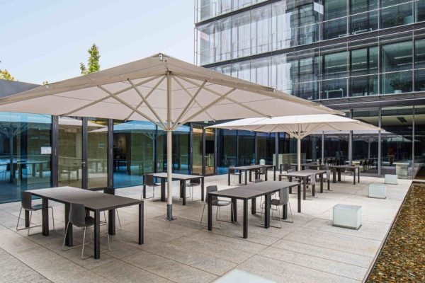 Weisse ALBATROS Grossschirme spenden Schatten auf einer modernen Firmen-Terrasse mit Tischen und Stühlen vor einem Glasgebäude.