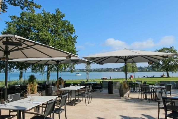 Elegante ALBATROS Grossschirme in Grau überdachen die Terrasse eines Restaurants direkt am See und bieten Gästen grosszügigen Schatten bei traumhafter Aussicht.
