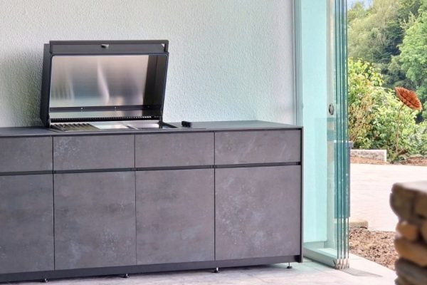 Avers Outdoor-Küche mit geöffnetem Grillmodul und eleganten dunklen Fronten in Steinoptik, platziert in einem modernen Outdoorbereich mit Glaswänden.