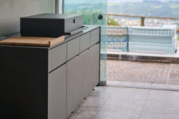 Moderne Avers Outdoor-Küche in dunkler Steinoptik mit Grillmodul und Holzarbeitsfläche, platziert in einem überdachten Outdoorbereich mit Blick auf die Landschaft.