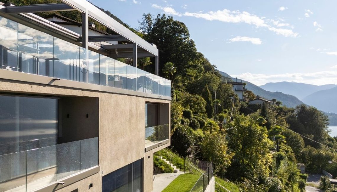 Seitliche Ansicht eines privaten Wohnhauses in Gerra im Tessin mit Lamellendach auf der Terrasse, Glasbrüstung und freiem Blick in die Hügellandschaft und Richtung See.