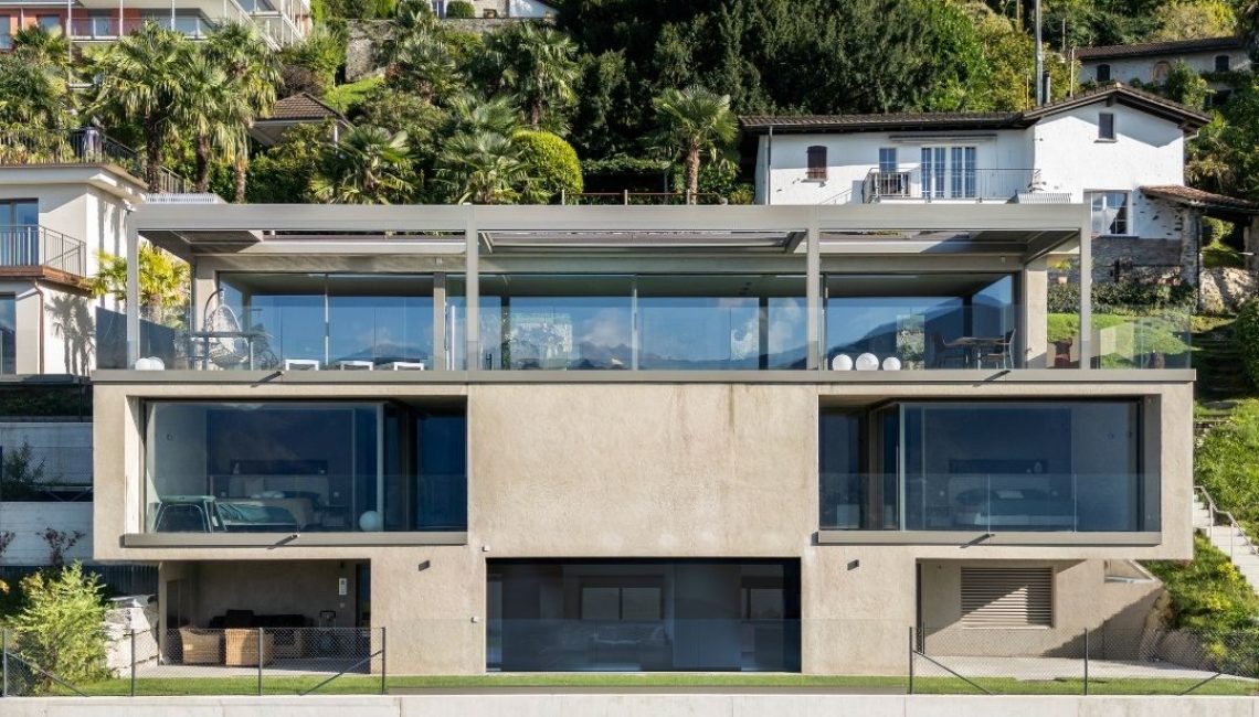 Frontansicht eines privaten Wohnhauses in Gerra im Tessin mit integriertem Lamellendach auf der oberen Terrasse und freier Sicht in die umliegende Landschaft.