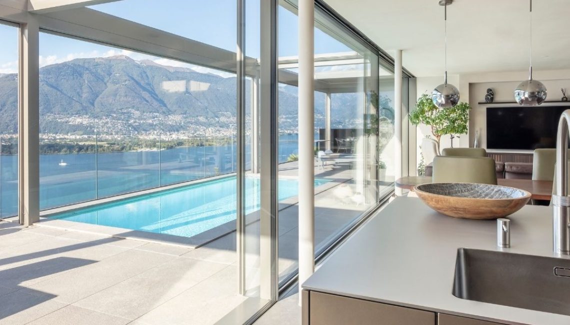 Grossflächige Glasfront mit Blick vom Wohnbereich auf die Terrasse mit Lamellendach, Pool und Seesicht in Gerra im Tessin.