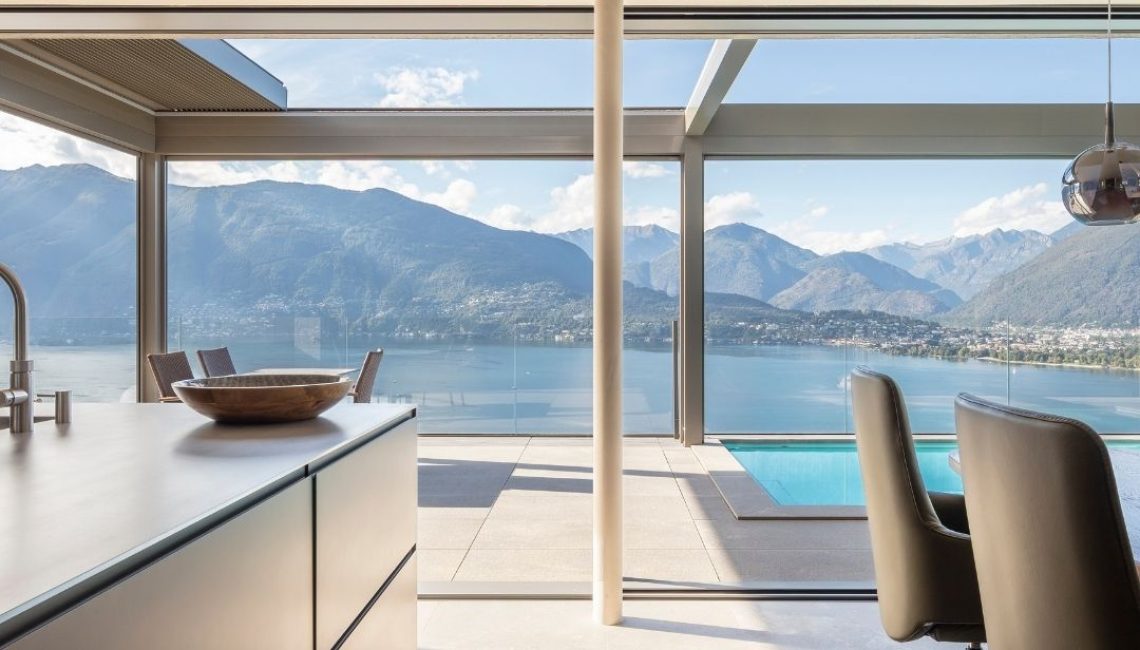 Blick aus dem Innenraum auf die Terrasse mit Lamellendach, Glasbrüstung und Pool mit freier Sicht auf den See und die umliegende Berglandschaft in Gerra im Tessin.