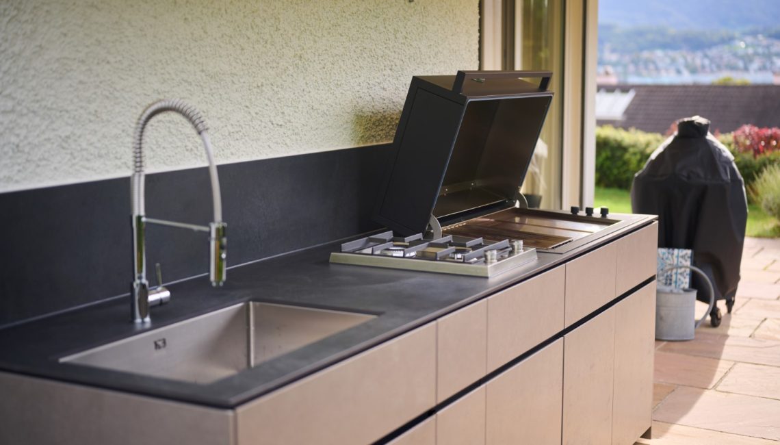 Aussenküche mit Spülbecken, Gasherd und geöffnetem Grill vor Keramik Rückwand
