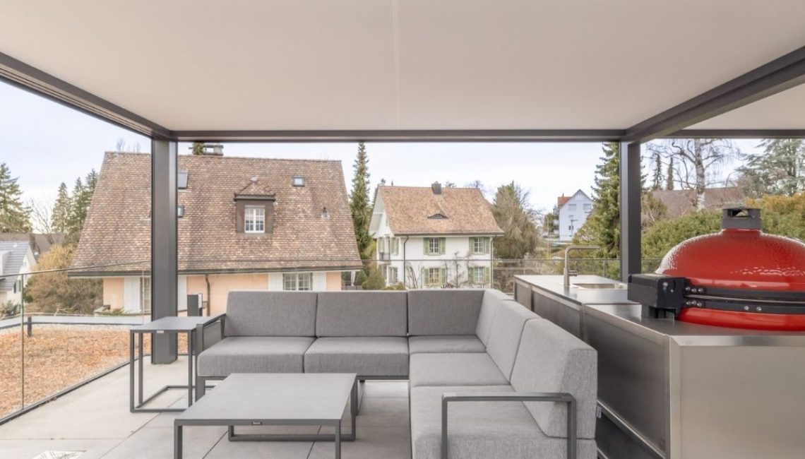 Überdachte Attikaterrasse mit grauem Lounge-Sofa und Outdoor-Küche unter einem Festdach ECL 130.