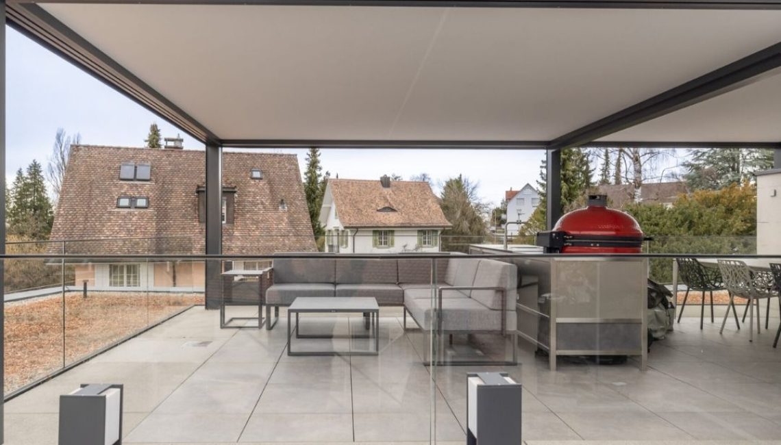 Modernes Festdach ECL 130 auf einer Attikaterrasse in Wallisellen mit Lounge, Glasgeländer und Outdoor-Küche mit rotem Grill.