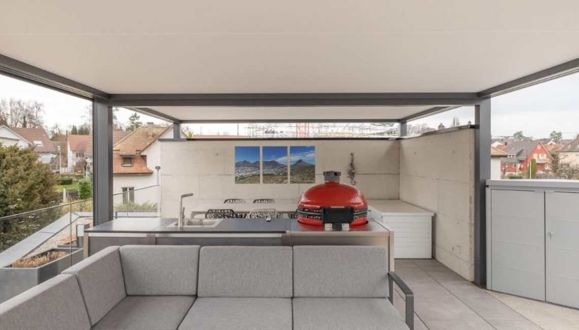 Moderne Outdoor-Lounge unter Festdach ECL 130 mit grauer Eckcouch und integrierter Outdoorküche samt rotem Grill.