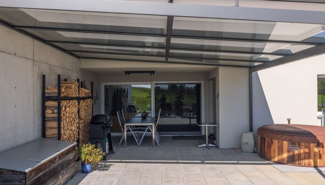 Terrasse mit modernem Glasdach – überdachter Aussenbereich mit Esstisch, Grill, Holzlager und Whirlpool.