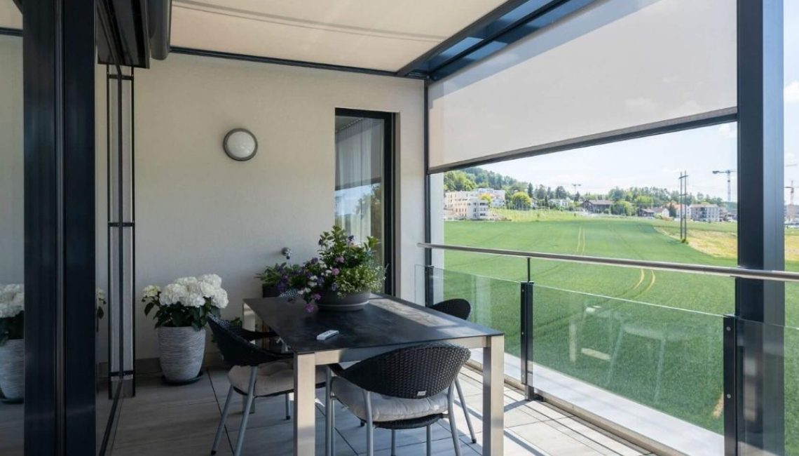 Überdachte Terrasse mit Glasdach und seitlichem Sonnenschutz in Uitikon Waldegg, modern möbliert mit Esstisch und Pflanzen.