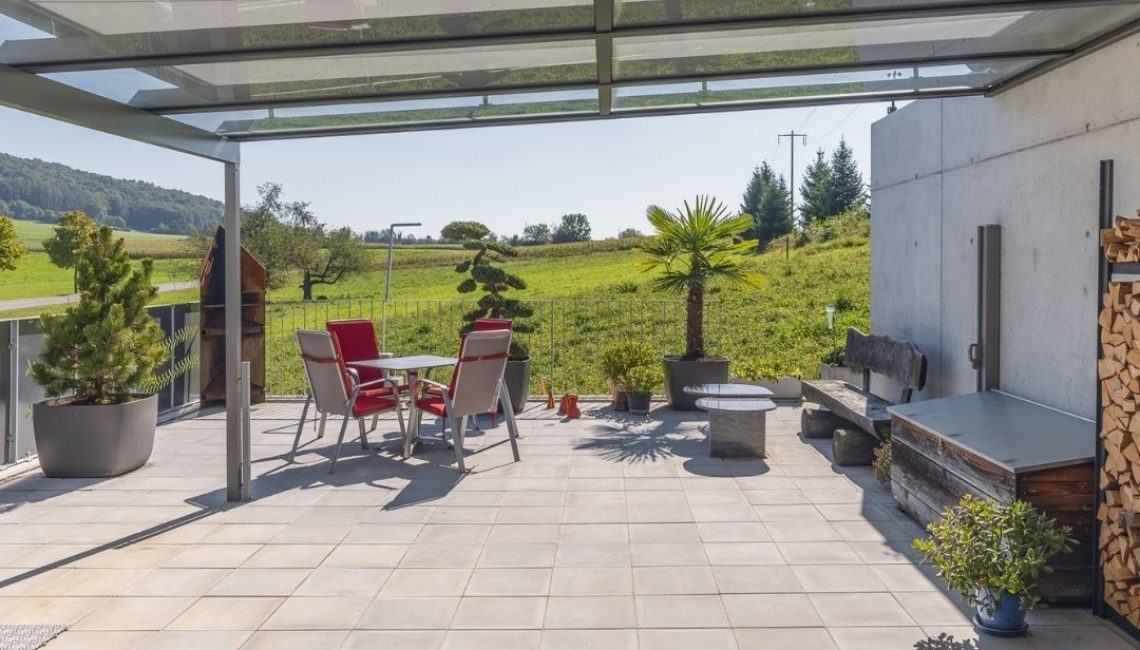 Überdachte Terrasse mit Glasdach – Sitzgruppe mit roten Stühlen, Pflanzen und weitem Ausblick ins Grüne.