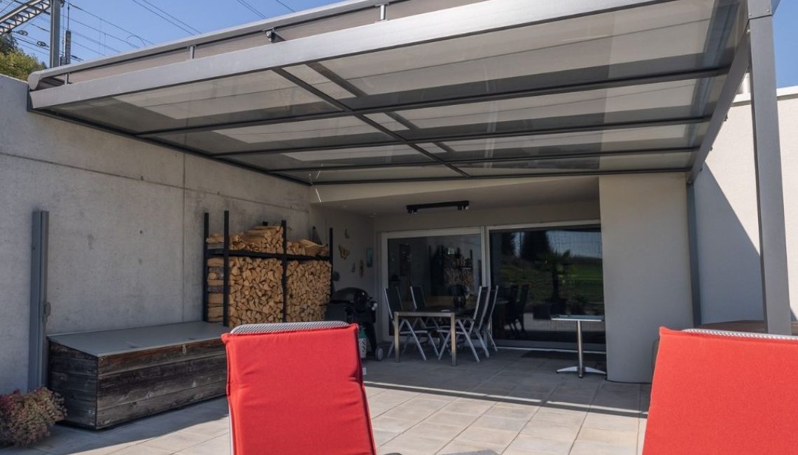 Terrasse mit modernem Glasdach – Blick vom Sitzbereich mit roten Stühlen auf den überdachten Essplatz und das Holzlager.