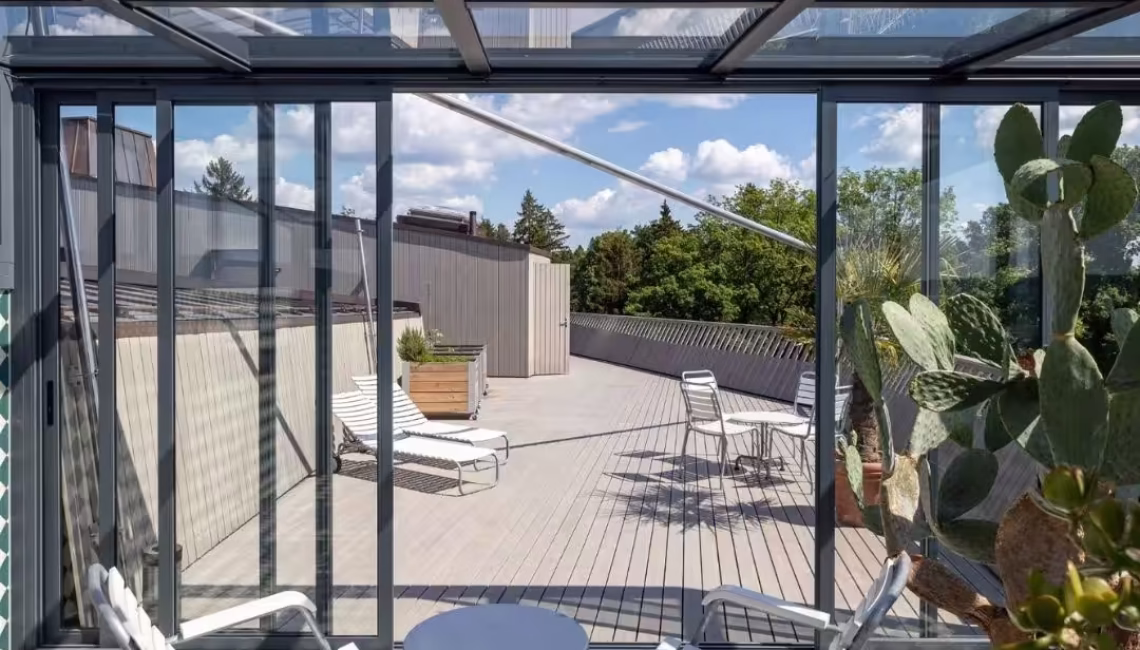 Innenansicht einer Glasdach Orangerie in Kloten mit Glaswänden, Dachverglasung und Blick auf die Dachterrasse mit Liegestühlen.