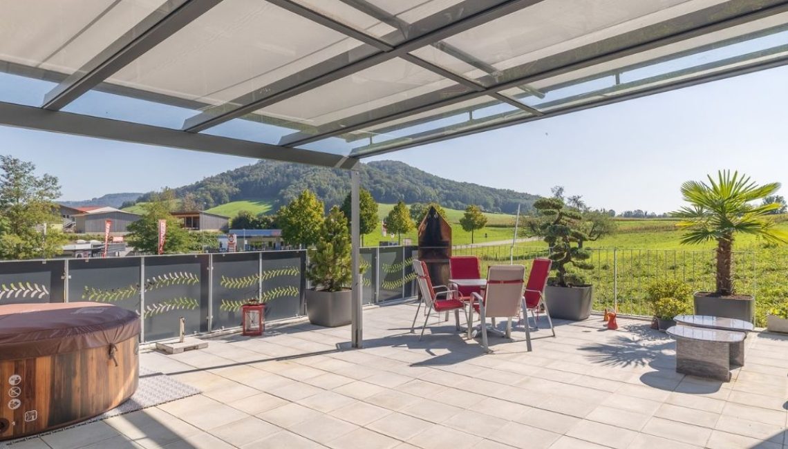 Terrasse mit Glasdach und Beschattung – Sitzgruppe mit roten Stühlen, Whirlpool und Pflanzen mit Ausblick auf grüne Hügel.