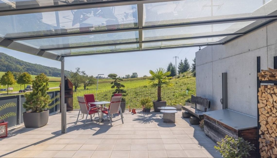 Grosszügige Terrasse mit Glasdach, modernen Gartenmöbeln und Blick ins Grüne – lichtdurchfluteter Outdoor-Bereich mit natürlichem Ambiente.