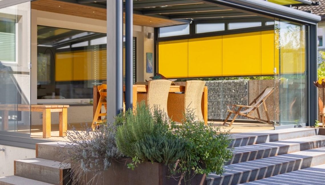 Moderner Wintergarten mit Glasdach, gelben Senkrechtmarkisen und Glasfronten, die sich vollständig öffnen lassen.