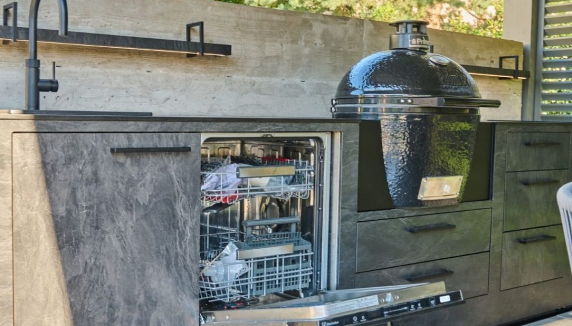 Outdoor-Küche aus Steinoptik mit integriertem Geschirrspüler, Spülbecken und Keramikgrill unter einem modernen Lamellendach.