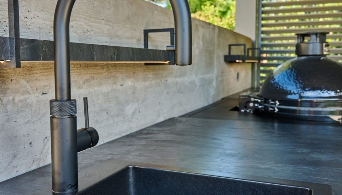 Detailaufnahme einer Outdoor-Küche mit schwarzem Spülbecken, moderner Armatur und Keramikgrill im Hintergrund.